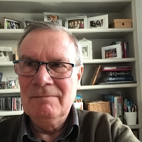 Older man with glasses taking a selfie in front of a bookcase filled with books, photos, and decorative items.