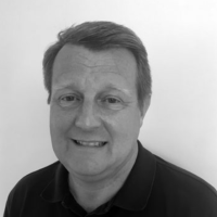 Man with short hair smiling, wearing a dark collared shirt, in front of a plain light background.