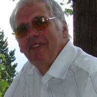 Older man in a white shirt and tinted glasses smiling outdoors near trees.