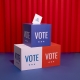 Ballot box with "VOTE" on it and a paper that says "GO VOTE" against a red curtain background.