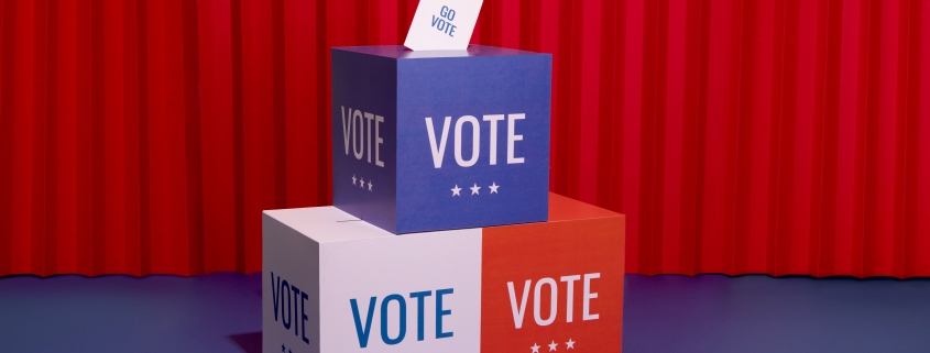 Ballot box with "VOTE" on it and a paper that says "GO VOTE" against a red curtain background.