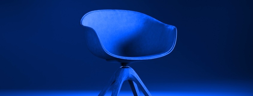 Modern, blue chair with wooden legs under dramatic blue lighting on a smooth surface.