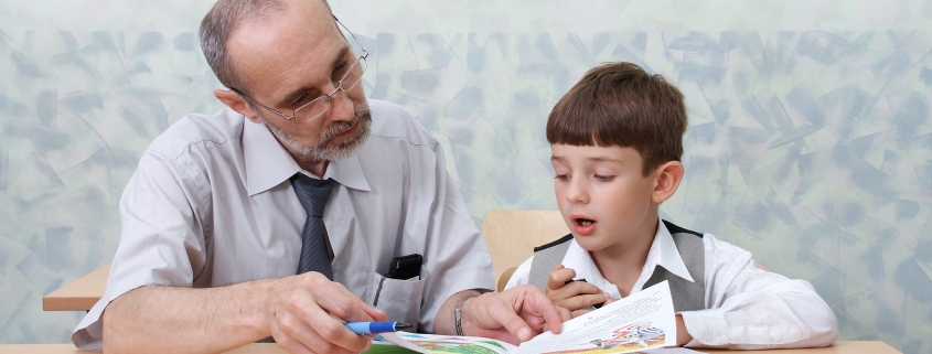 An older man helps a young boy read a book at a desk in a classroom setting.