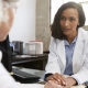 A female doctor sits at a desk, holding a patient's hand and listening attentively in an office setting.