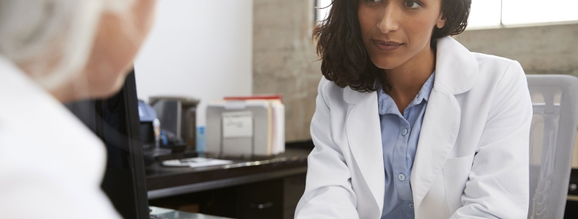 A female doctor sits at a desk, holding a patient's hand and listening attentively in an office setting.
