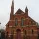 Red brick church with arched windows, two tall spires, and a central red door on a cloudy day.