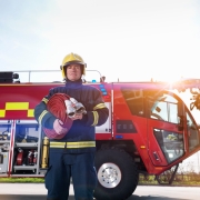 Firefighter in uniform holding a fire hose, standing in front of a large red fire engine outdoors.