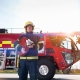 Firefighter in uniform holding a fire hose, standing in front of a large red fire engine outdoors.