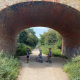 Three children with bikes stand under a brick bridge on a sunny, tree-lined dirt track.