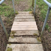 Wooden footbridge with metal railings over grass, featuring two new planks among older, mossy boards.