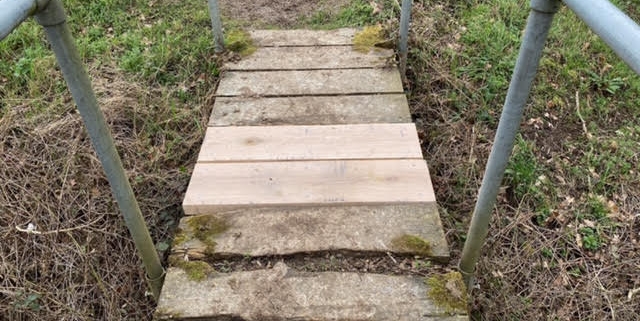 Wooden footbridge with metal railings over grass, featuring two new planks among older, mossy boards.