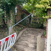 Concrete steps lead up to a green gate, with trees and a building nearby; a barrier blocks the lower steps.