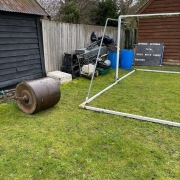 Roller and metal frame on grass near wooden sheds, fence, barrels, scoreboard, and garden tools in background.
