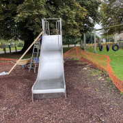 A metal slide under construction, with a ladder beside it and orange safety fencing around the area.
