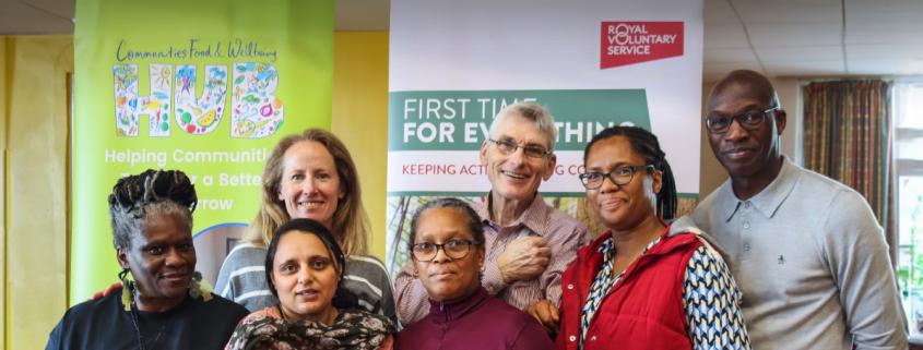 A diverse group of seven adults smiling together at a community event, with banners in the background.
