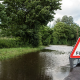 A road is flooded with water, and a warning sign with "Flood" stands at the edge of the water.