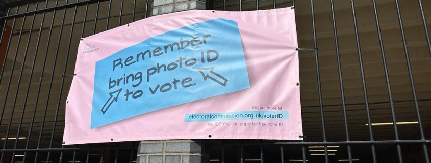 Pink banner on a fence reads: "Remember bring photo ID to vote" with blue arrows pointing to the text.