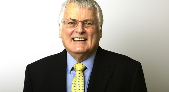 Smiling older man in a black suit, blue shirt, and yellow tie against a plain light background.