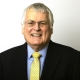 Smiling older man in a black suit, blue shirt, and yellow tie against a plain light background.