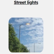 A street light is on during the day, with trees and a partly cloudy sky in the background.