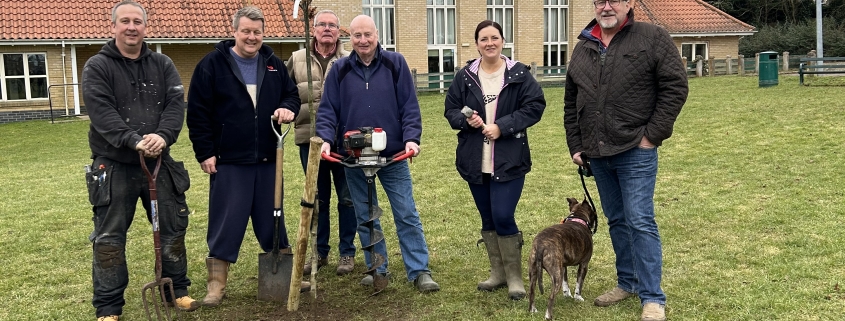Six people and a dog pose with spades by a newly planted tree on a grassy area in front of a building.