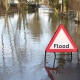 Flooded road with a triangular warning sign that reads “Flood” in the foreground.