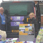 Two smiling people at an info stall with leaflets and pamphlets beside a van outside a building.