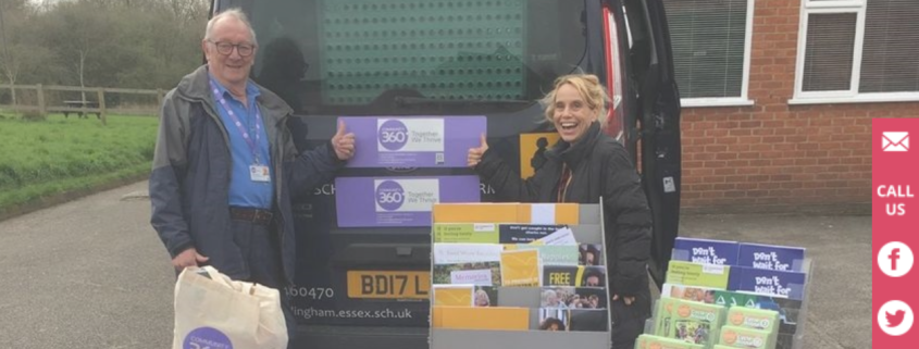 Two smiling people at an info stall with leaflets and pamphlets beside a van outside a building.