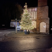 A Christmas tree with lights and baubles stands by a brick building on a wet street at night.