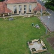 Aerial view of a building with a red roof, playground, garden beds, grass lawn, and nearby car park.