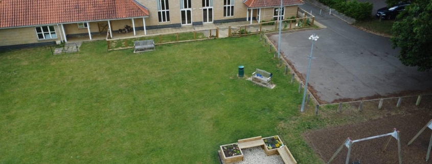 Aerial view of a building with a red roof, playground, garden beds, grass lawn, and nearby car park.