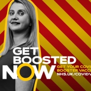 Woman stands by "Get Boosted Now" COVID-19 booster vaccine message on a red and yellow striped background.
