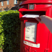 A hand posts a letter in a red Royal Mail priority postbox on a sunny day.
