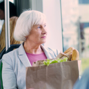 Older woman holding groceries sits on a bus, looking out the window with a thoughtful expression.