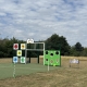 Outdoor playground with basketball hoop, numbered blocks, and green target wall on grass under a partly cloudy sky.